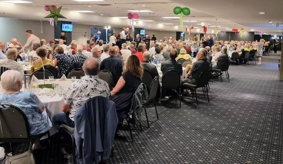 A large indoor gathering of attendees seated at tables during a gala dinner, with decorations and a stage in the background.