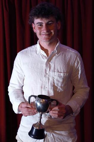A young man wearing a white shirt, smiling while holding a trophy in front of a red curtain.